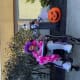 A young adult sits in her wheelchair wearing a purple wig with a bright pink feather boa next to Halloween decorations.