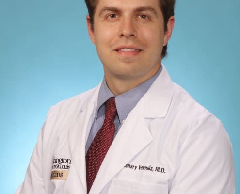 Dr. Vesoulis in a white lab coast, short wavy hair and a red tie smiles for his professional headshot.