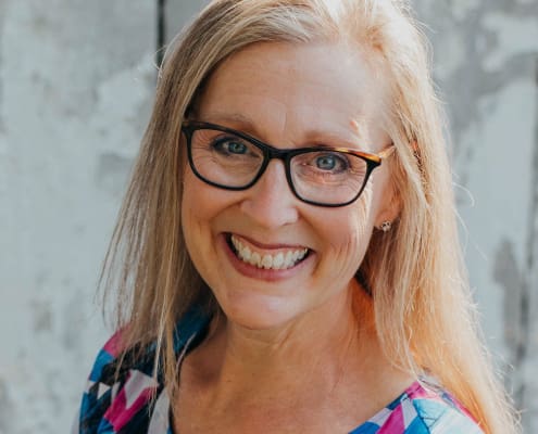 A woman smiling broadly with long blond hair, dark round glasses and a multi-color blouse.
