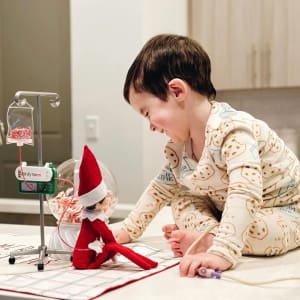 A boy with cerebral palsy smiles at his red elf on a shelf.