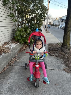 A toddler with cerebral palsy smiles on her red trike out on the sidewalk.
