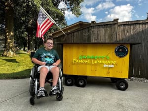 A young man in a wheelchair sits next to a neon sign advertisting his lemonade