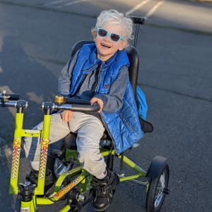 A boy with cerebral palsy and white hair smiles broadly out on his adaptive trike.