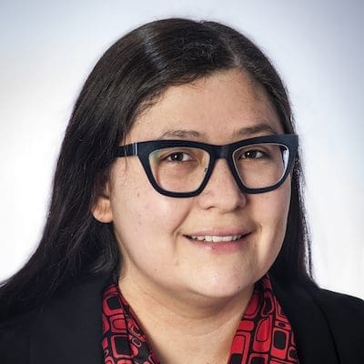 Dr Fuentes, a woman with long brown hair parted on one side and dark rimmed glass smiles in a black blazer and red shirt.