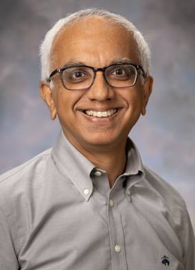 A man with short white hair and glasses smiles in a gray button down shirt.