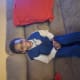A young boy in blue pant suit smiles sitting on a couch.