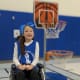 A young girl with long dark hair in her wheelchair by a basketball net.