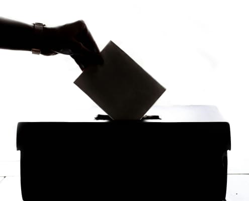 A black and white image of a hand slipping a vote into a ballot box.