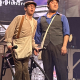 Two young men wearing flat caps acting out a scene on stage with one having cerebral palsy standing in his walker