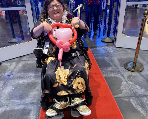 A woman in a wheelchair smiling in her dress and a tiara and holding a pink heart shaped balloon